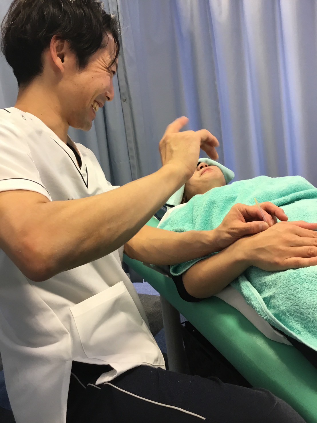 施術者が笑顔で患者様の手元に鍼施術を行っている様子。合谷や足三里など、花粉症に関係するツボを通じて自律神経や免疫バランスを整える施術風景。